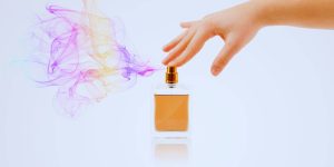 Mão feminina acionando o spray de um frasco de perfume, que solta uma nuvem aromática colorida