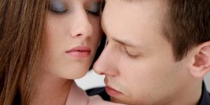 Close nos rostos de um casal de olhos fechados; o homem parece estar sentido o perfume da mulher