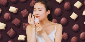 Mulher oriental cheirando um frasco de perfume; ao fundo, uma montagem com chocolates