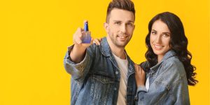 Casal sorridente abraçado; homem mostra um frasco de perfume