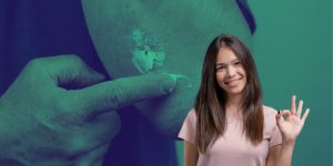 Pessoa passando pomada no cotovelo ao fundo e mulher jovem fazendo sinal de aprovação à frente